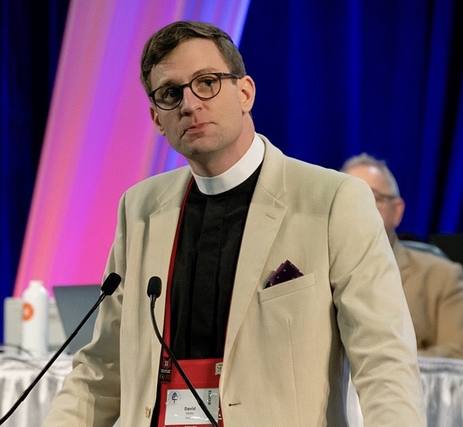 Photo of David Sibley, wearing suit and clerical collar at a lectern.